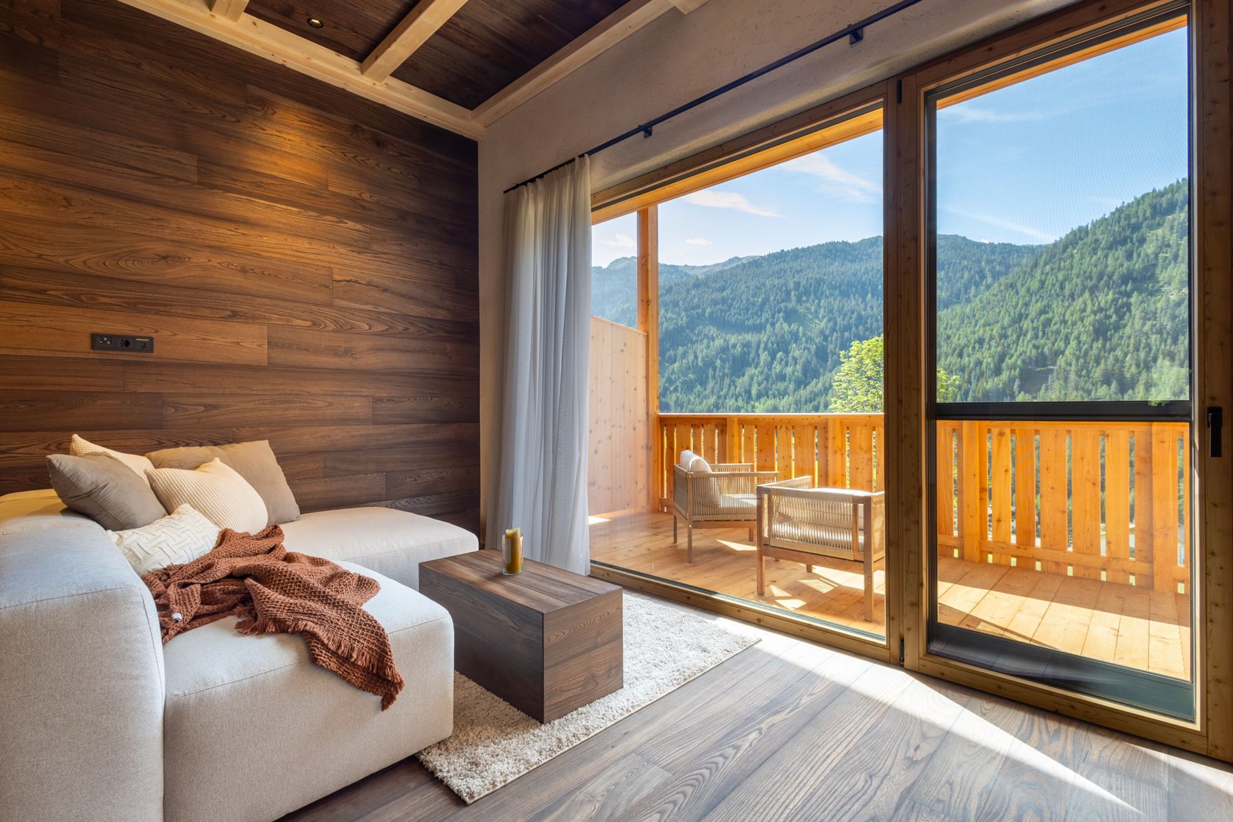 Gemütliche Holzstube mit Sofa und Blick vom Balkon auf die bewaldeten Berge des Sarntals